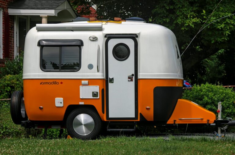 A bright orange and white Armadillo trailer parked in a suburban yard, surrounded by greenery.